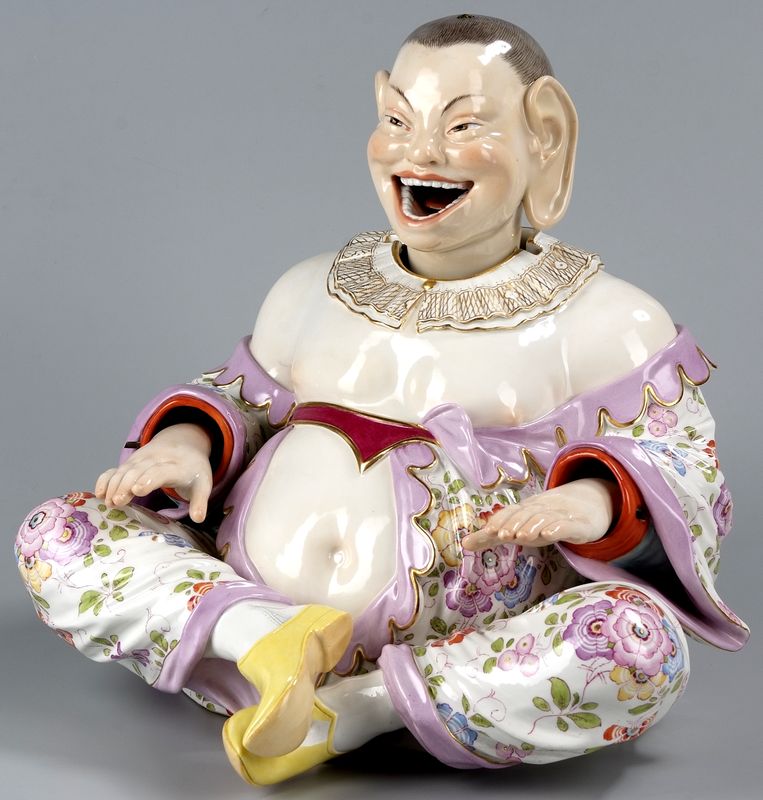 Porcelanowa postać Chińczyka siedzącego po turecku. Biało-fioletowy strój. Jego zewnętrzne warstwy zdobione wzorem kwiatowym. Twarz roześmiana. Oczy skośne i przymróżone, usta szeroko otwarte. Bardzo duże uszy.