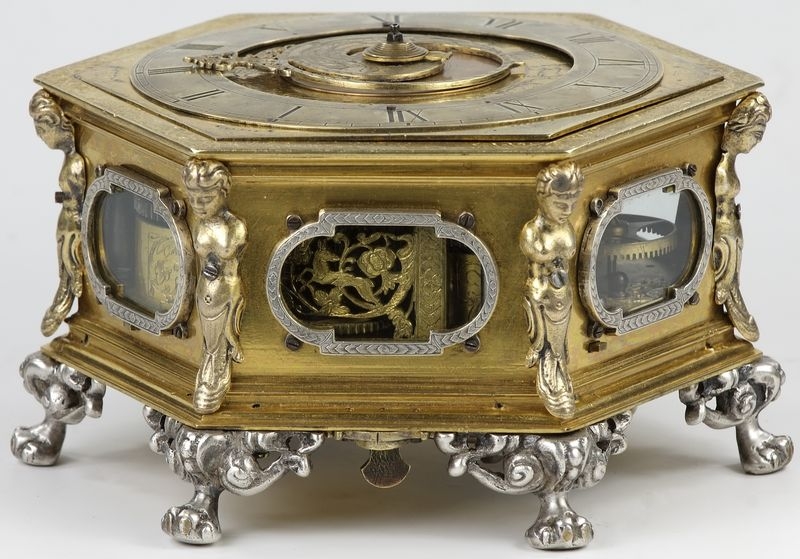Złoty zegar przypominający formą szkatułkę. Nóżki srebrne. U szczytu, położona płasko na pudełku tarcza zegarowa. 