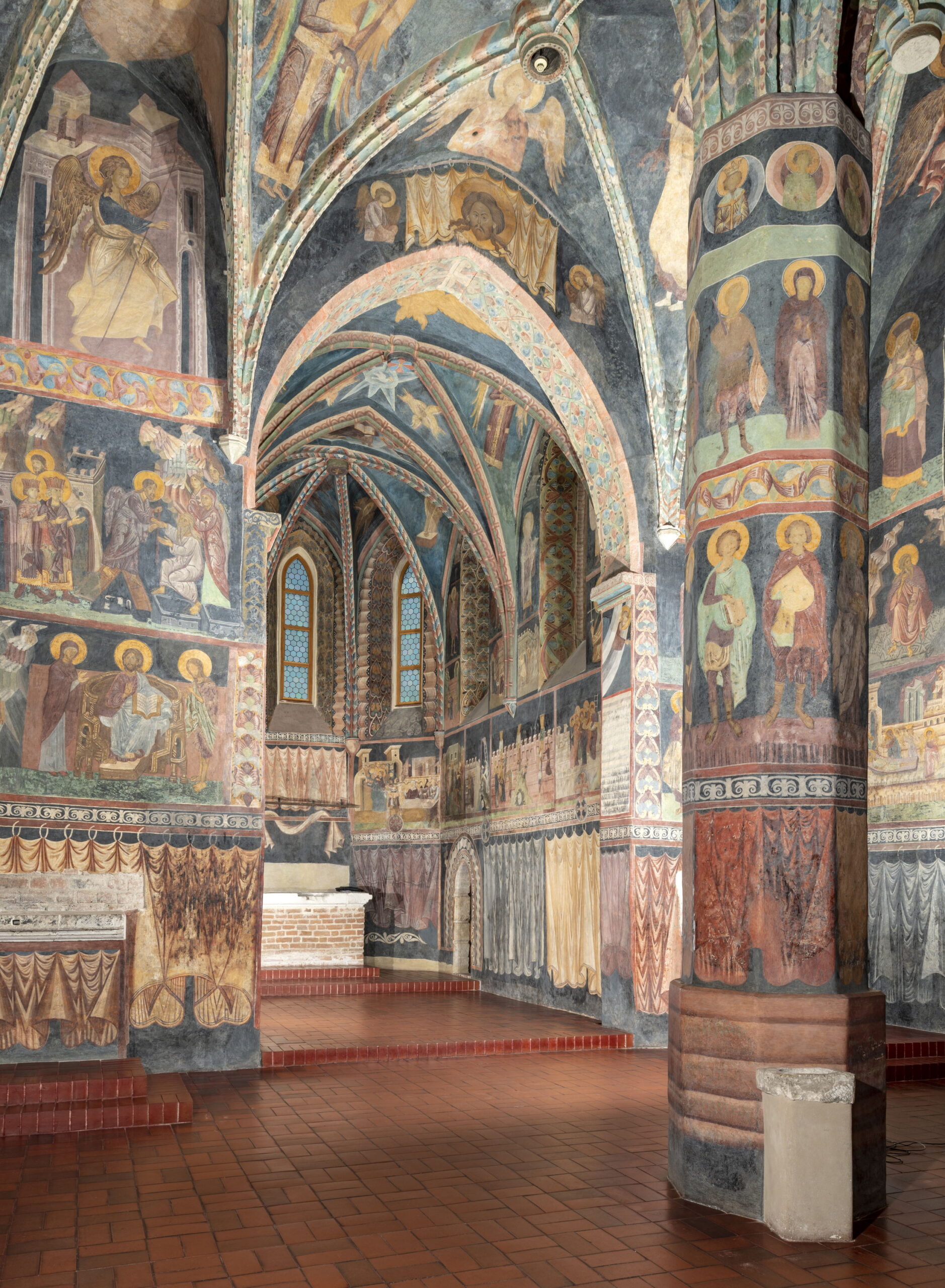 Fotografia wnętrza kaplicy. Ceglana podłoga. Po prawej filar podtrzymujący sklepienie. Ściany i sufit ozdobione kolorowymi malowidłami o tematyce religijnej.