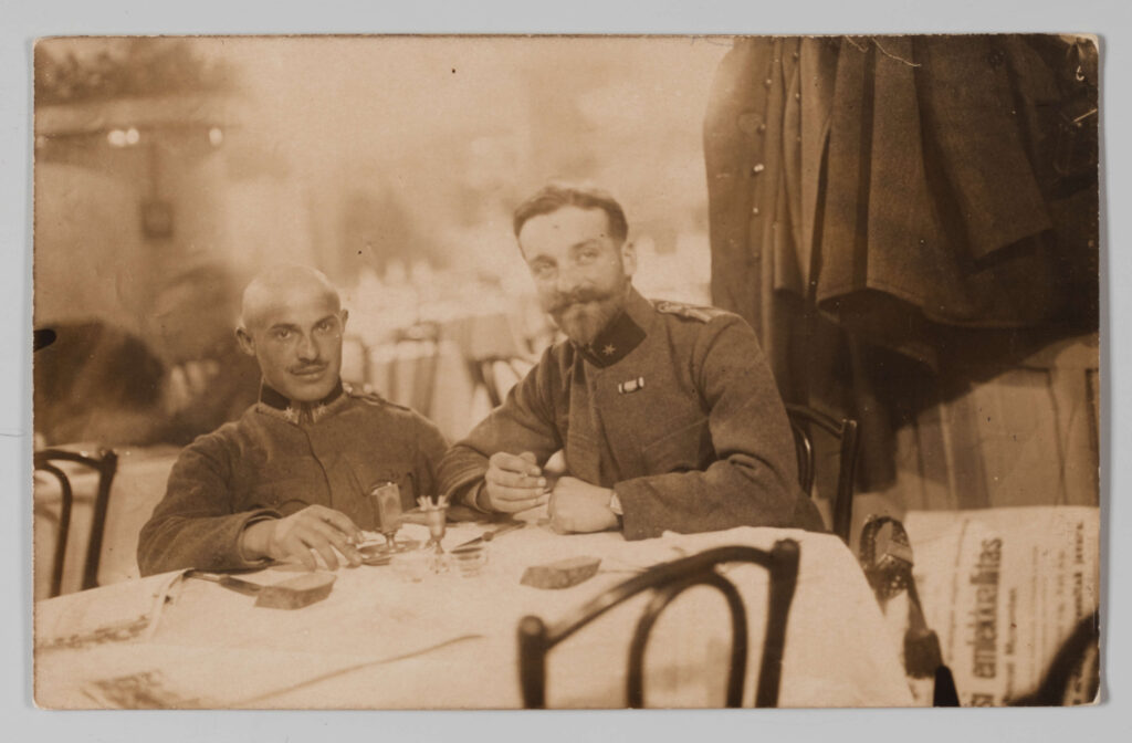 Fotografia przedstawia dwóch mężczyzn siedzących przy stoliku w kawiarni, ubranych w mundury wojskowe. Z prawej strony Jerzy Pol z długimi wąsami i brodą. Obok niego kolega z ogoloną głową i krótko przystrzyżonymi wąsami. W tle widoczne wnętrze kawiarni ze stolikami nakrytymi białymi obrusami.