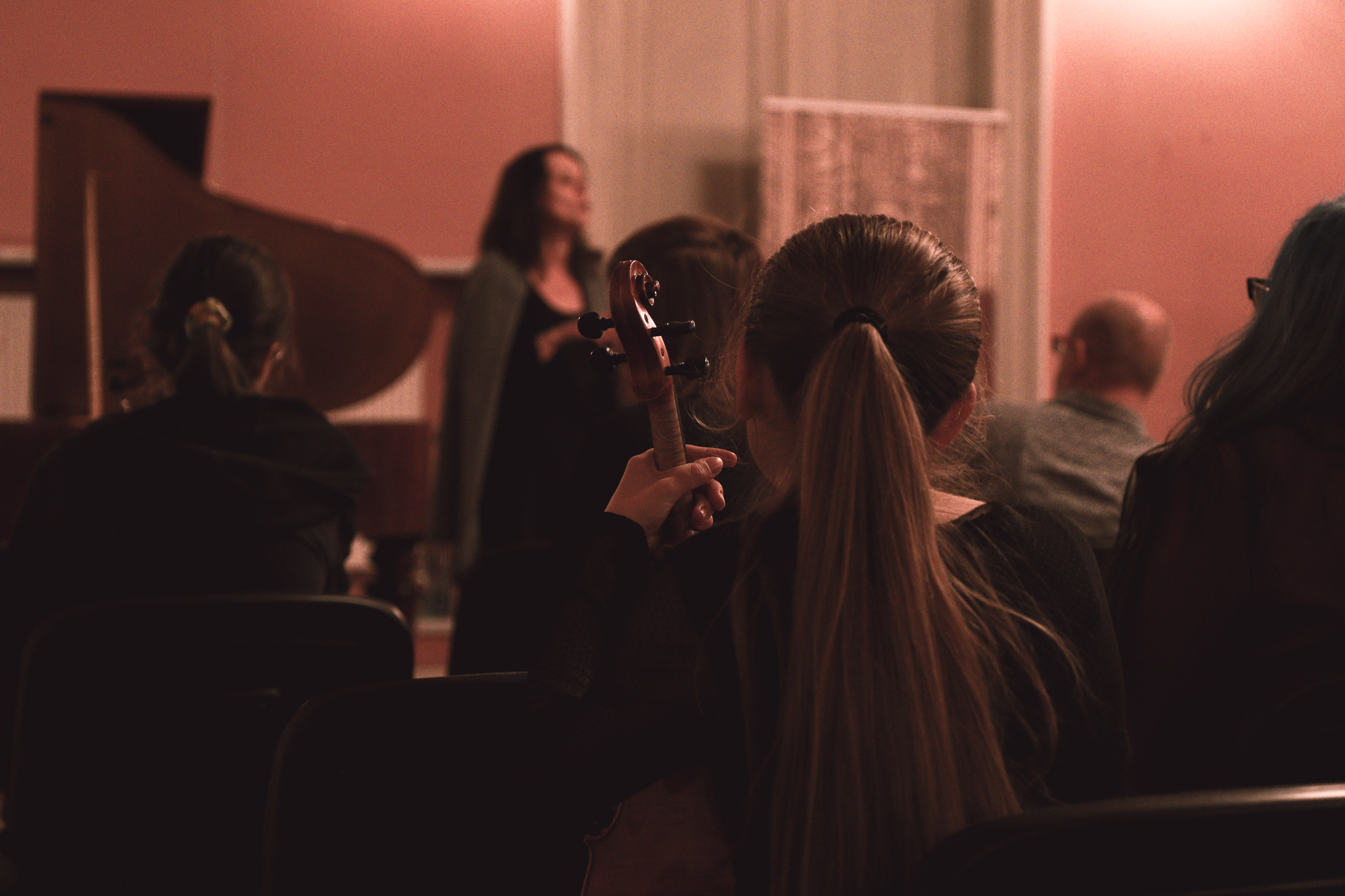 Na pierwszym planie siedzi dziewczyna z długimi włosami. W dłoniach trzyma skrzypce. Widoczna od tyłu. W tle widać kobietę zapowiadającą koncert.