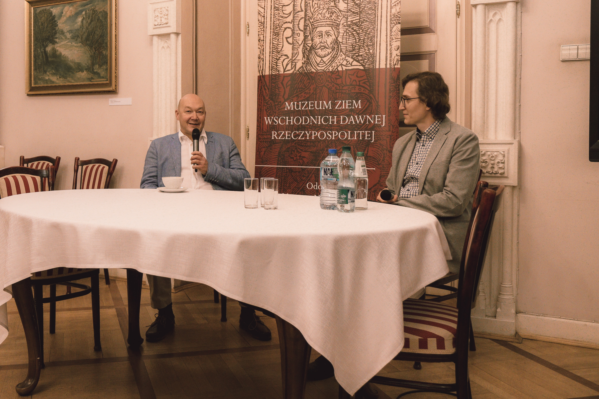 Dwóch mężczyzn siedzi przy stole. Po lewej Wojciech Orliński - autor książek o Stanisławie Lemie. Trzyma w dłoni mikrofon. Po prawej doktor Przemysław Kaliszuk, który prowadzi spotkanie z pisarzem.