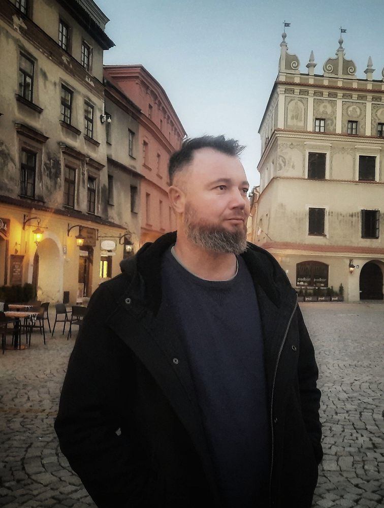 mężczyzna krótko ścięty z zarostem na czarno ubrany w tle kamienice starego miasta w Lublinie