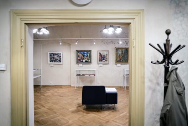 Fragment sali wystawowej. Widoczne są obrazy wiszące na ścianach, stojące gabloty z eksponatami i fotel na środku pomieszczenia. Na pierwszym planie po prawej stronie znajduje się płaszcz na wieszaku.
